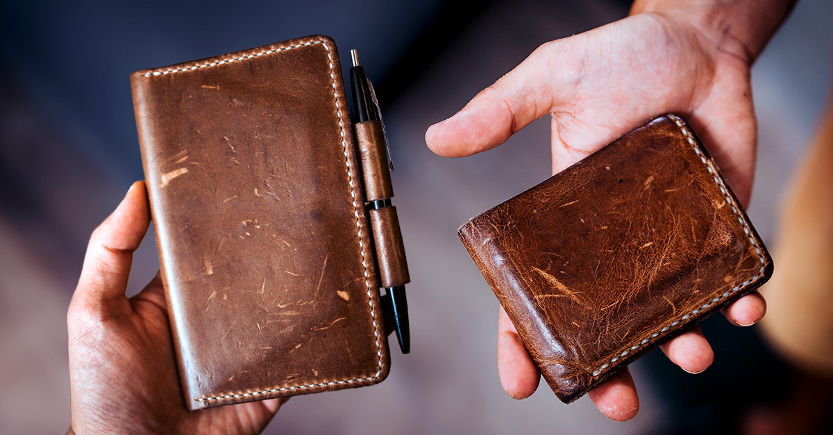 Two hands; one holding a cracked leather A5 notebook and the other holding a cracked leather wallet