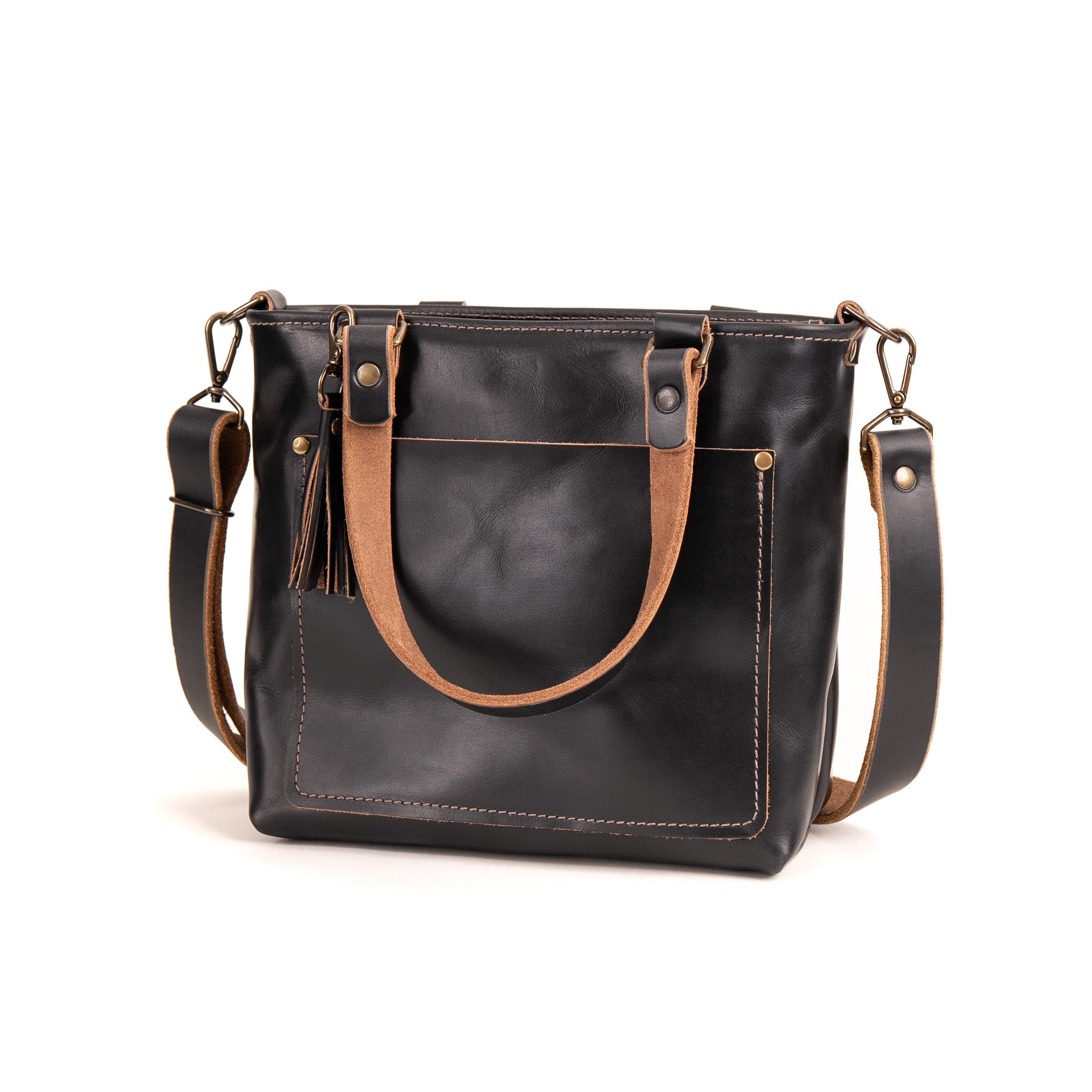 A black leather tote bag with tan leather handles and trim, featuring a front pocket with a tassel detail and a detachable shoulder strap, shown on a plain white background.