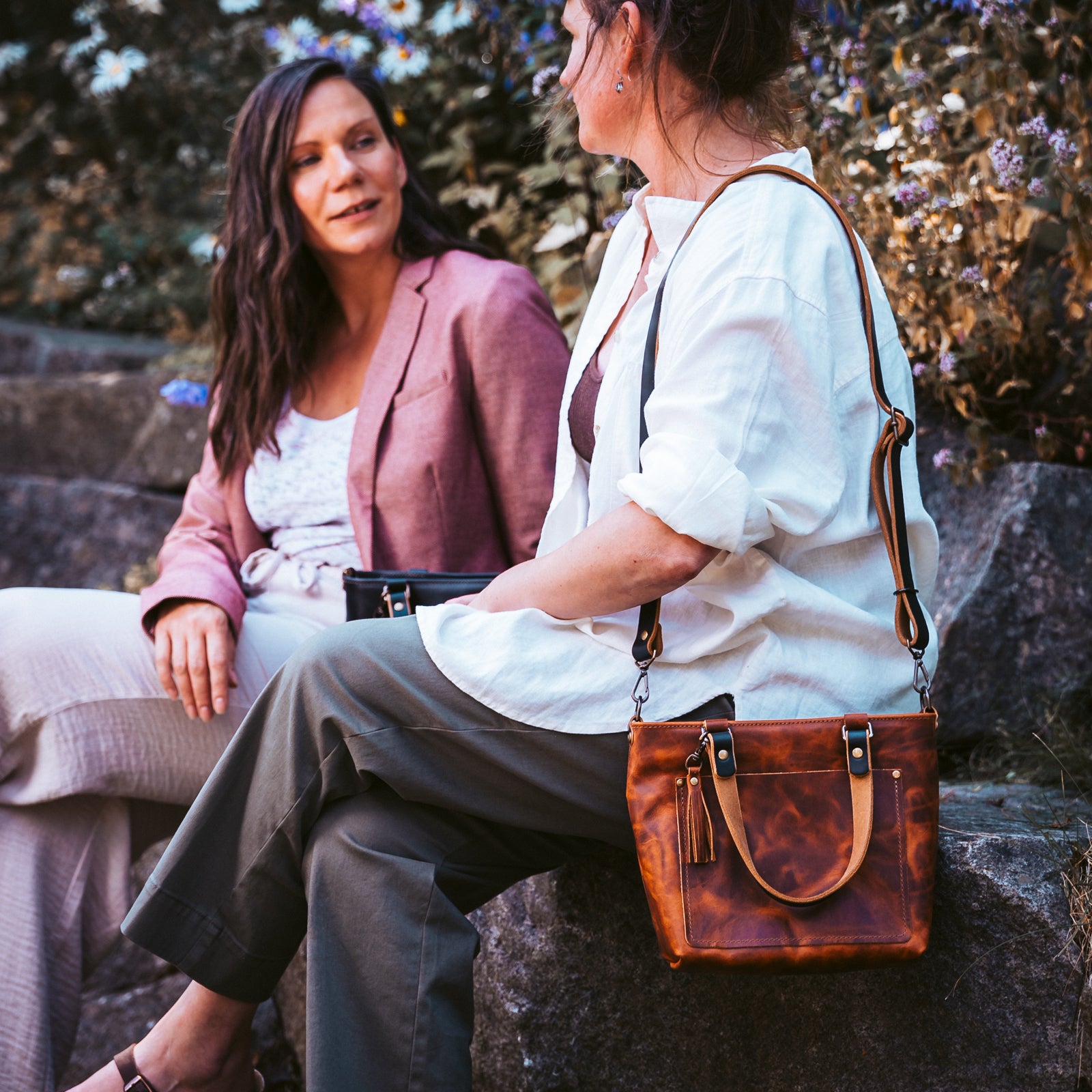 A woman in a white blouse and gray trousers sits outdoors on a stone bench, wearing a brown leather crossbody handbag with a tassel and front strap. Another woman in a pink blazer sits beside her, holding a black clutch.