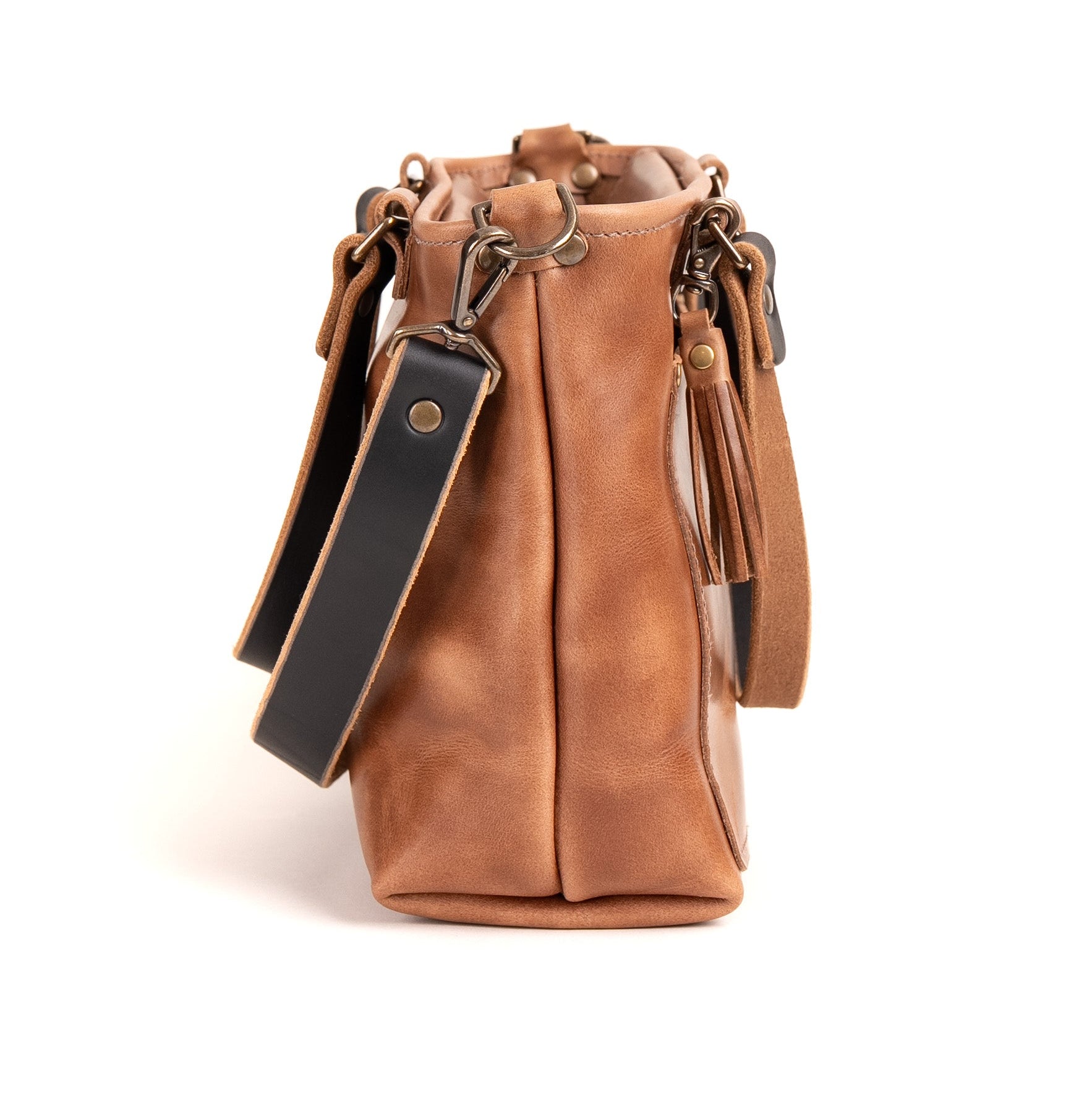 A tan leather bucket bag with a black leather strap, gold-tone hardware, and tassel details, shown against a plain white background.